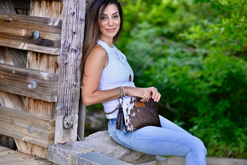 K&ouml;nnte beinhalten: Eine Frau in einem wei&szlig;en Tanktop und blauen Jeans h&auml;lt eine braune und wei&szlig;e Clutch aus Kuhfell mit einem braunen Quastenanh&auml;nger.