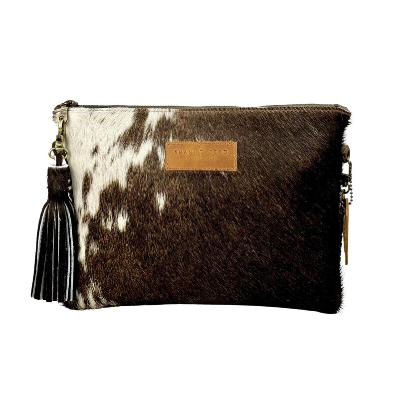 K&ouml;nnte beinhalten: Eine braune und wei&szlig;e Clutch aus Kuhfell mit einem braunen Leder-Quaste und einem braunen Leder-Etikett mit der Aufschrift "CHAND TWEED".
