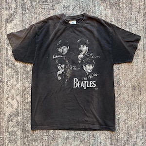 May include: A charcoal gray t-shirt featuring a black and white image of The Beatles. The image includes the band members' faces and signatures, with "The Beatles" printed below. The shirt has a vintage, worn look.