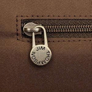 May include: Close-up of a brown fabric with a zipper and a silver-colored zipper pull. The pull is circular and has the words "JIM THOMPSON" engraved on it. The zipper is dark brown.