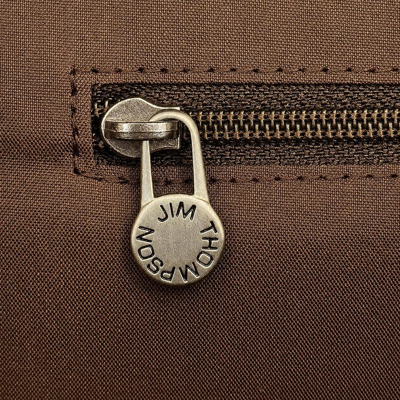 May include: Close-up of a brown fabric with a zipper and a silver-colored zipper pull. The pull is circular and has the words "JIM THOMPSON" engraved on it. The zipper is dark brown.