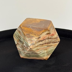May include: A geometric polyhedral stone carving featuring banded layers of brown, tan, cream, and sage green mineral deposits. The object has a faceted, multi-sided shape with a smooth, polished surface that highlights the natural horizontal striations.