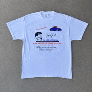 May include: A white short-sleeve t-shirt featuring a graphic for the Fifth Annual Car Show Extraordinaire held on September 3rd, 2001. The design includes a blue and red car, a caricature of Jerry Lewis, and text referencing the MDA Labor Day Telethon.