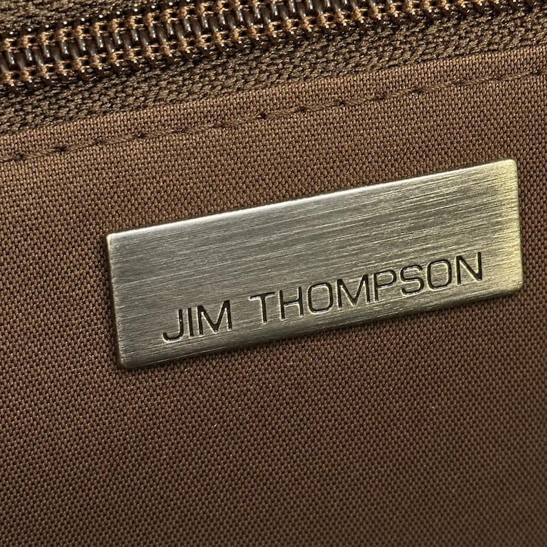 May include: Close-up of a brown fabric with a zipper and a rectangular metal tag. The tag is brushed silver and reads "JIM THOMPSON" in capital letters. The fabric appears to be a textured weave.