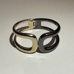Puede incluir: Un brazalete de metal dorado y negro con un diseño simple y moderno. El brazalete está abierto y tiene un cierre de bisagra.