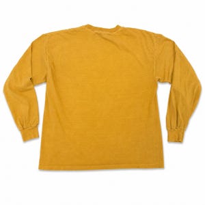 Puede incluir: Camiseta de manga larga de color amarillo mostaza. La prenda tiene cuello redondo y mangas largas con pu&ntilde;os acanalados. La tela parece ser un material suave y ligeramente descolorido. La camiseta es de color liso, sin gr&aacute;ficos ni patrones visibles.