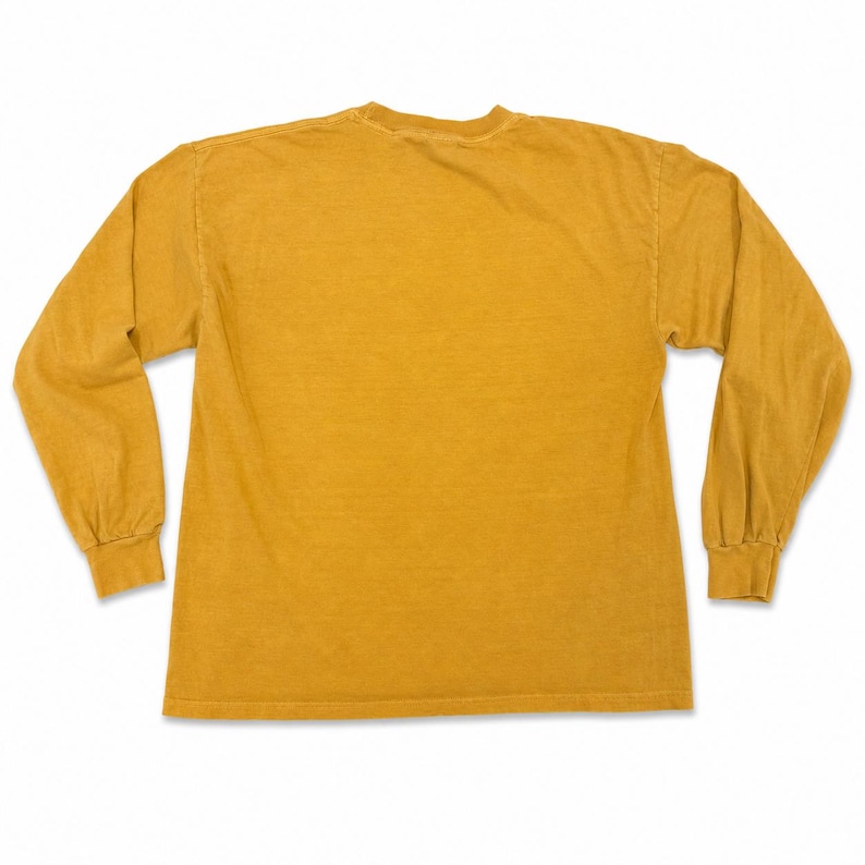 Puede incluir: Camiseta de manga larga de color amarillo mostaza. La prenda tiene cuello redondo y mangas largas con pu&ntilde;os acanalados. La tela parece ser un material suave y ligeramente descolorido. La camiseta es de color liso, sin gr&aacute;ficos ni patrones visibles.