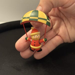 Pode incluir: Uma pequena figura de madeira de uma personagem com fato e gorro vermelhos com rebordo branco, suspensa por fios vermelhos de um paraquedas aos quadrados amarelos e verdes. A personagem tem uma cara pintada com bochechas rosadas.