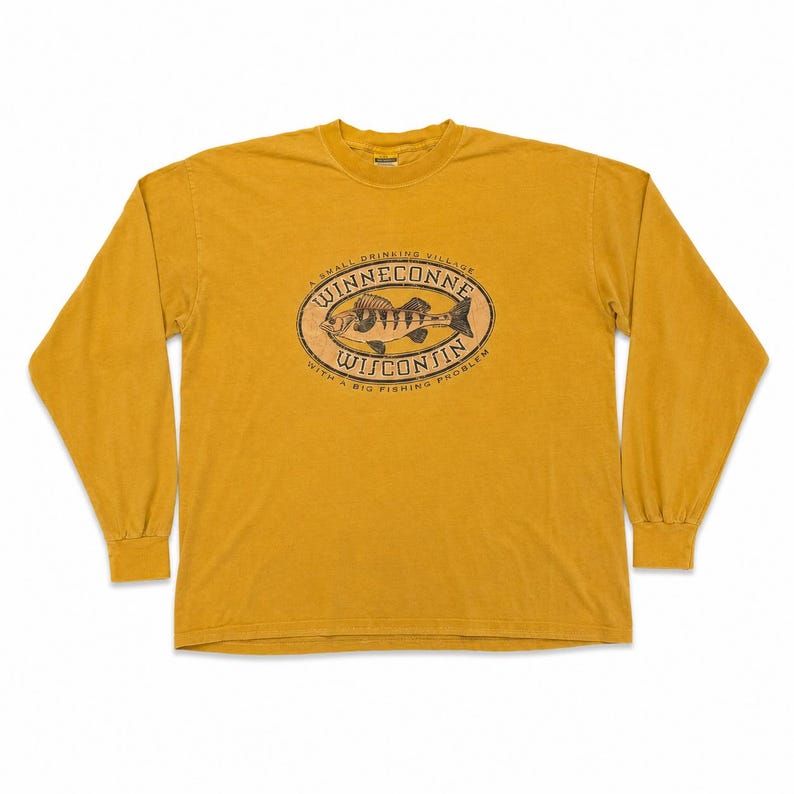 Puede incluir: Camiseta de manga larga de color amarillo mostaza con un gr&aacute;fico en la parte delantera. El gr&aacute;fico es un dise&ntilde;o ovalado con una ilustraci&oacute;n de pez y las palabras "WINNECONNE WISCONSIN" y "A SMALL DRINKING VILLAGE WITH A BIG FISHING PROBLEM".