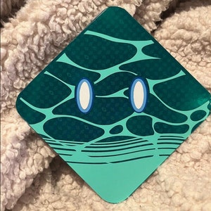 Puede incluir: Una pegatina en forma de diamante de color verde azulado y verde oscuro con un diseño de dibujos animados. La pegatina presenta dos ojos ovalados blancos con contornos azules y un patrón de líneas interconectadas y pequeños puntos.