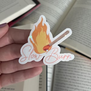 May include: A white sticker with a drawing of a lit match and the text "Slow Burn" in pink cursive. The sticker has a glittery finish.