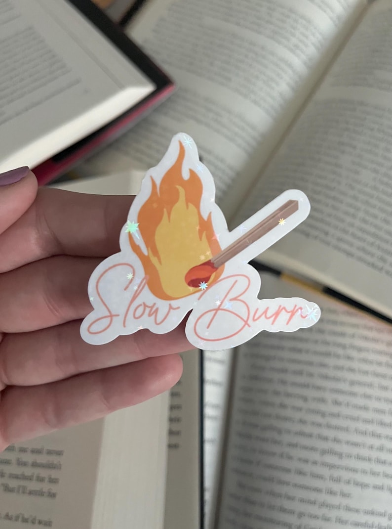 May include: A white sticker with a drawing of a lit match and the text "Slow Burn" in pink cursive. The sticker has a glittery finish.