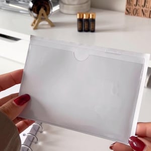 Op de afbeelding: Een doorzichtige plastic zak met een witte rand, ontworpen om papieren of kaarten in te bewaren. De zak wordt in een hand gehouden.