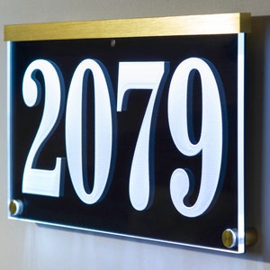 May include: A black acrylic house number sign with gold trim and white numbers. The number is 2079.