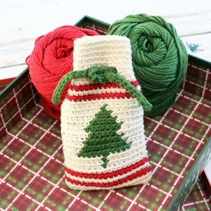 May include: A white crocheted drawstring bag with red and green stripes and a green crocheted Christmas tree design. The bag is sitting in a box with a red and green plaid pattern. Two balls of yarn, one red and one green, are also in the box.