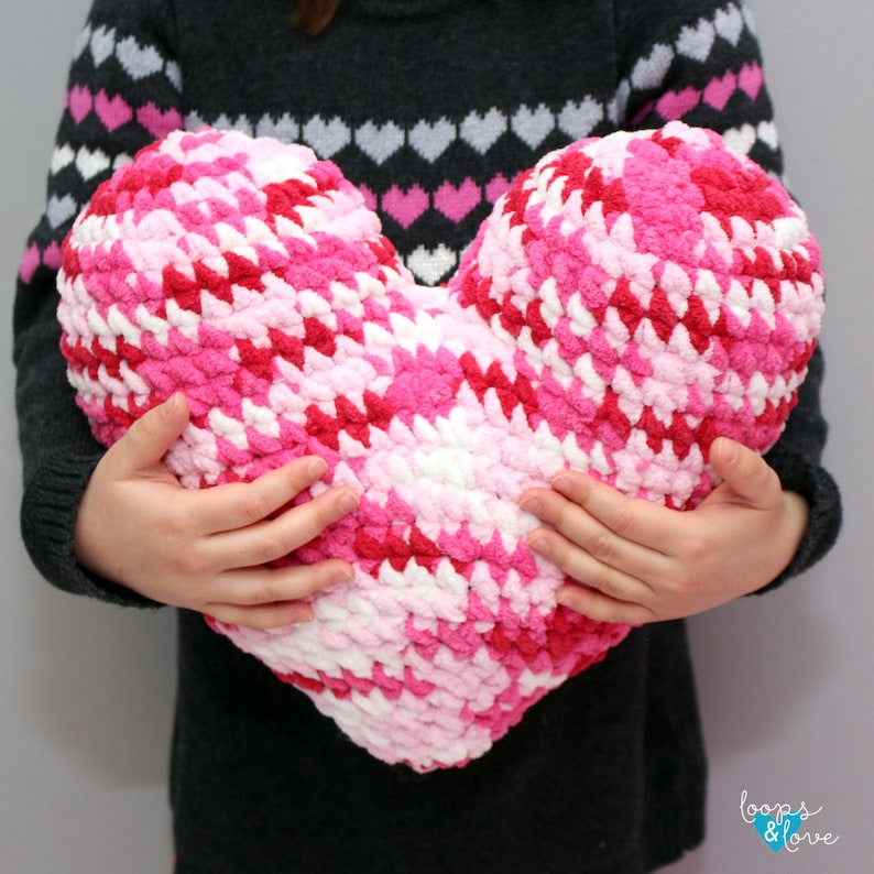 May include: A large pink, red, and white crocheted heart-shaped pillow. The pillow is being held by a person.