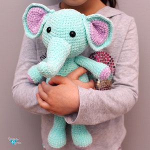 Peut inclure: Un éléphant en peluche crocheté bleu clair avec des oreilles violettes. L'éléphant a des yeux noirs en forme de boutons et une longue trompe recourbée.