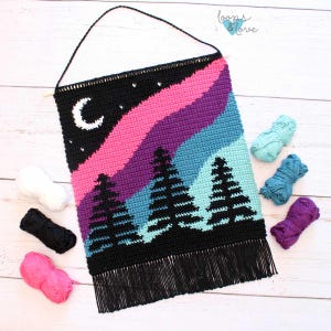 May include: A black crochet wall hanging featuring a colourful aurora borealis design and three black silhouette trees. The hanging has a fringe border and is made with different coloured yarn.