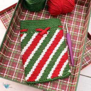 May include: A green and red striped crocheted bag with a white yarn bow. The bag is sitting in a plaid box with green and red yarn balls and a crochet hook.