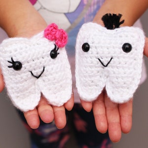 May include: Two crocheted tooth plush toys, one with a pink bow and the other with a black hair tuft. Both have black eyes and a smiling mouth.