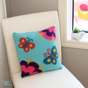 May include: A blue crocheted pillow with a colorful butterfly design. The butterflies are pink, red, yellow, and blue. The pillow is sitting on a white chair.