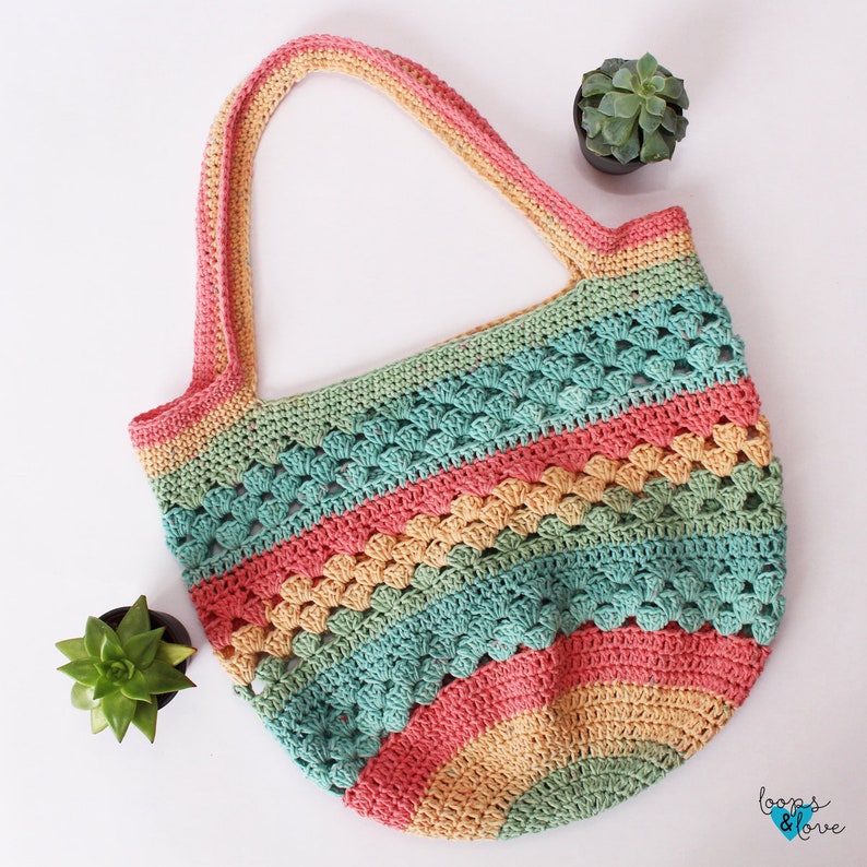 May include: A colourful crocheted tote bag with a rainbow pattern in shades of pink, green, and yellow. The bag has a wide, open top and two long handles.