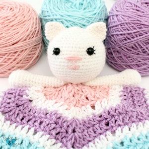 May include: A crocheted white cat with black eyes and a pink nose is attached to a multi-colored crocheted blanket. The blanket is made of pink, white, purple, and blue yarn.