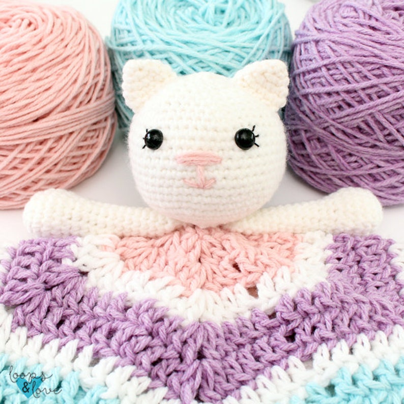 May include: A crocheted white cat with black eyes and a pink nose is attached to a multi-colored crocheted blanket. The blanket is made of pink, white, purple, and blue yarn.