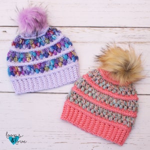 May include: Two crocheted hats with colourful yarn and faux fur pom poms. The hats are on a white wooden background. The hats are both in a bobble stitch pattern. One hat is purple and the other is pink.