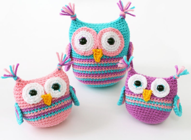 May include: Three crocheted owl plushies in different sizes. The largest owl is turquoise with pink and purple stripes. The medium owl is pink with purple and turquoise stripes. The smallest owl is purple with turquoise and pink stripes.