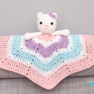 May include: A crocheted baby blanket with a pink, white, light blue, and purple chevron pattern. A white crocheted cat with a purple flower on its head is attached to the blanket.