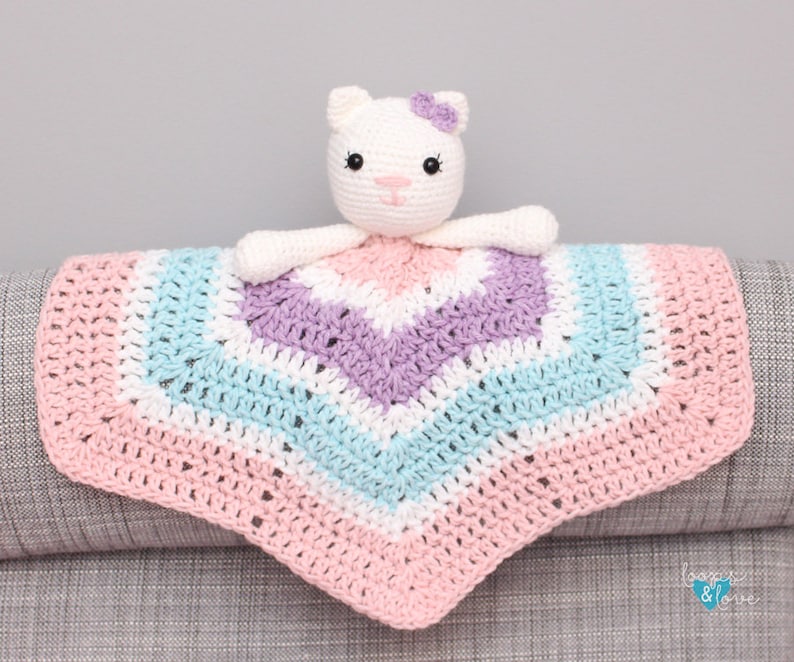 May include: A crocheted baby blanket with a pink, white, light blue, and purple chevron pattern. A white crocheted cat with a purple flower on its head is attached to the blanket.