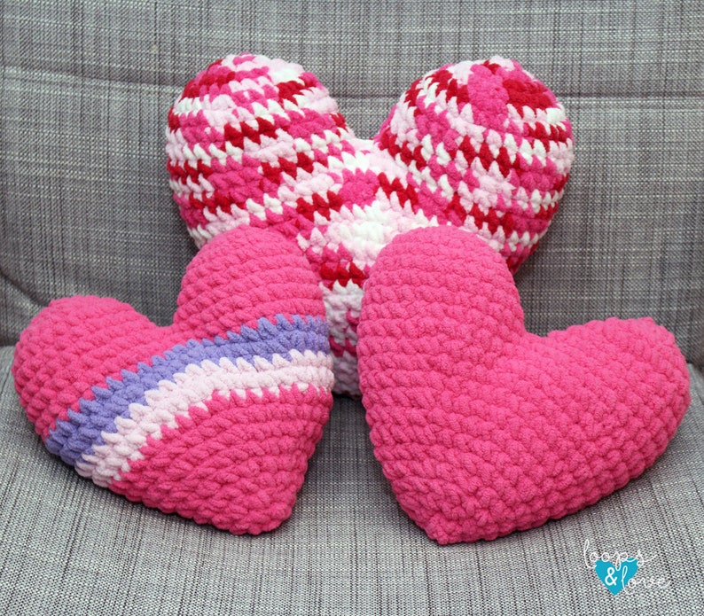 May include: Three crocheted heart-shaped pillows in pink, white, and purple. The pillows are arranged on a gray fabric surface. The text "Loops & Love" is visible in the bottom right corner of the image.