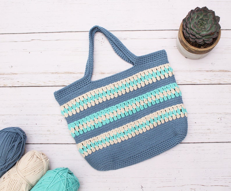 May include: A blue crocheted tote bag with white and turquoise stripes. The bag has a wide, flat base and two handles.