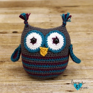 May include: A crocheted owl toy with gray, teal, purple, white, and yellow yarn. The owl has large eyes with black pupils and white and purple accents. The owl has a small yellow beak.