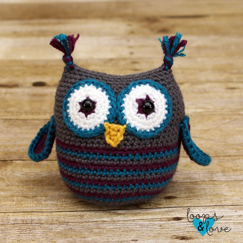 May include: A crocheted owl toy with gray, teal, purple, white, and yellow yarn. The owl has large eyes with black pupils and white and purple accents. The owl has a small yellow beak.