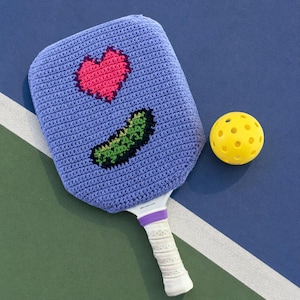 Op de afbeelding: Een lichtpaarse gehaakte hoes voor een pickleball-racket met een roze hart en een groen augurkontwerp, vergezeld van een felgele geperforeerde pickleball. Het racket heeft een wit handvat met een paarse gripband aan de basis van het blad.