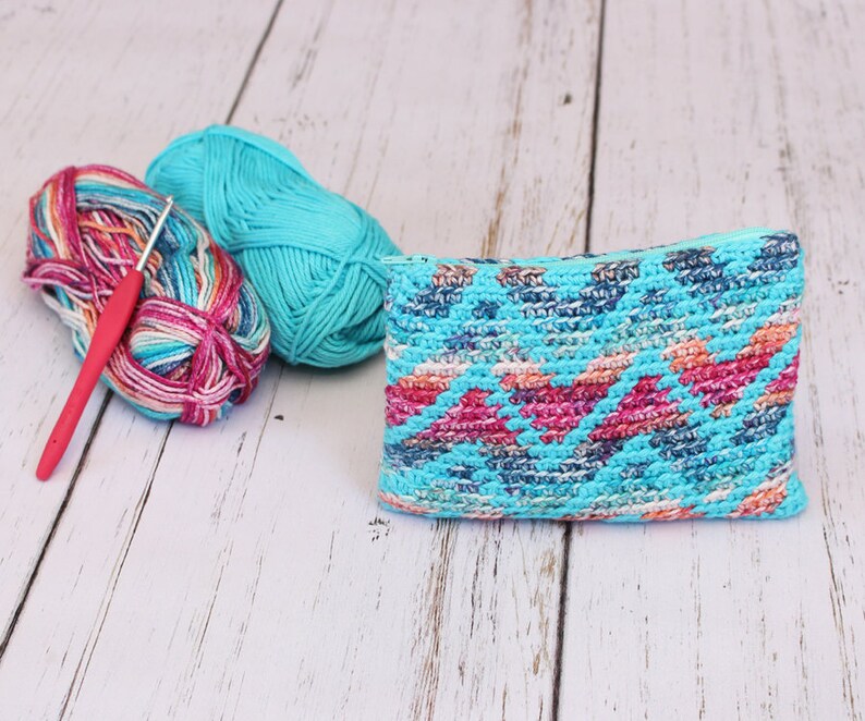 Puede incluir: Una bolsa de crochet turquesa con un patr&oacute;n geom&eacute;trico colorido, dos ovillos de lana y un ganchillo.