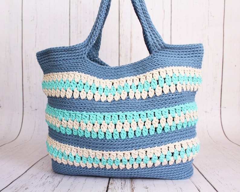 May include: A blue crocheted tote bag with white and turquoise stripes. The bag has a wide, flat base and two long handles.