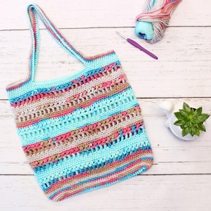 May include: A handmade crochet tote bag in shades of blue, pink, and tan. The bag features a striped pattern and long handles. A ball of yarn, a crochet hook, and a small succulent in a white pot are also visible.