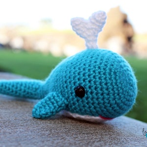 Puede incluir: Un juguete de ballena azul de crochet con una aleta blanca de crochet. La ballena tiene un pequeño ojo negro y una boca roja.