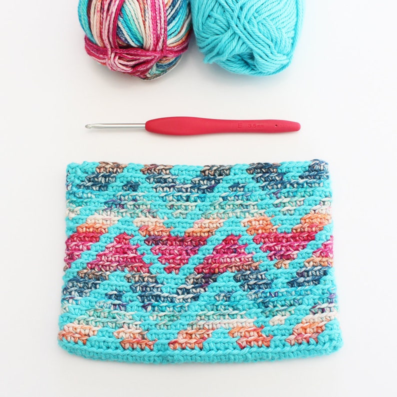 Puede incluir: Una diadema de crochet con un patr&oacute;n geom&eacute;trico en tonos turquesa, rosa y blanco. La diadema est&aacute; hecha con un hilo grueso y tiene un aspecto texturizado.
