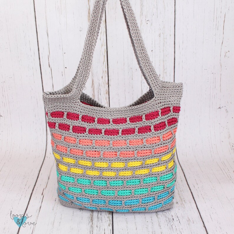 May include: A gray crochet tote bag with rainbow colored stripes. The bag has a wide, flat base and two long straps.