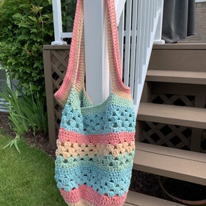 May include: A crocheted tote bag with a rainbow colour pattern of pink, yellow, green, and blue. The bag is hanging on a white railing.