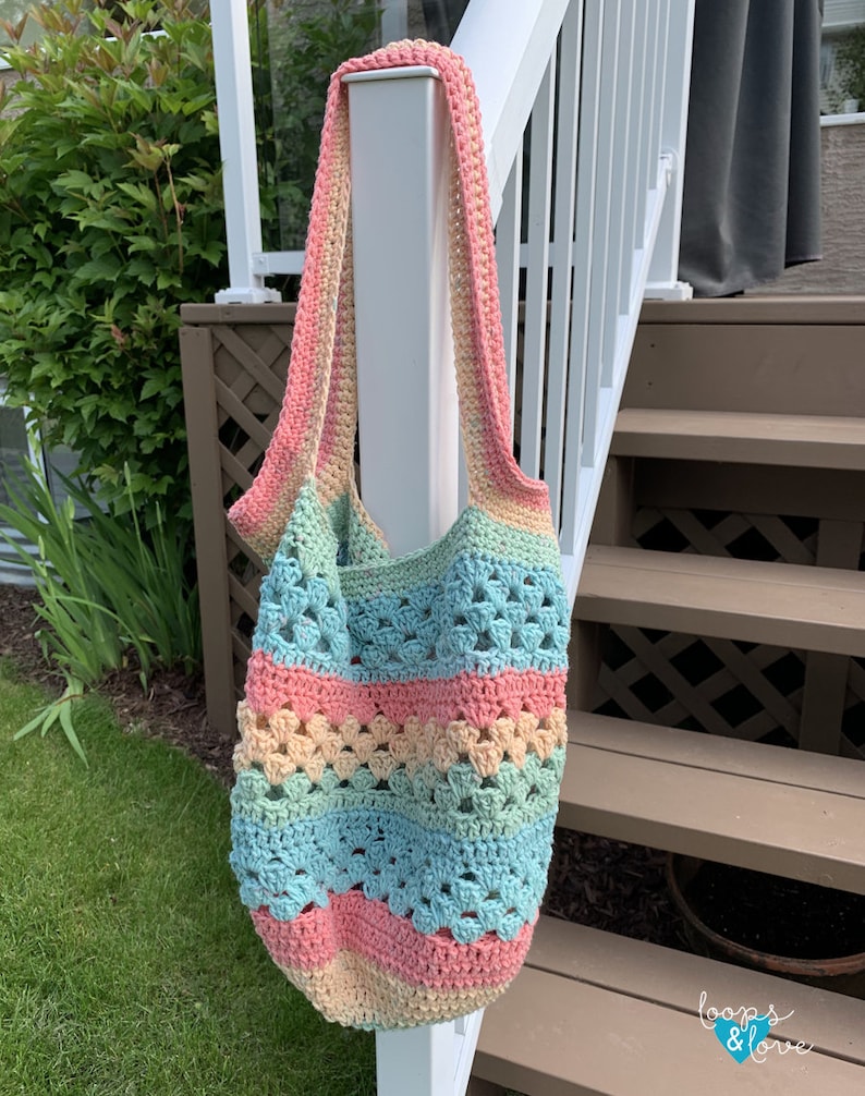 May include: A crocheted tote bag with a rainbow colour pattern of pink, yellow, green, and blue. The bag is hanging on a white railing.