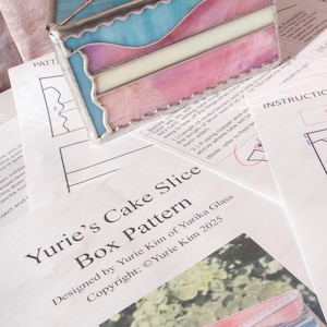 May include: A stained glass cake slice box with a red cherry on top. The box features layers of blue, pink, and white glass, outlined in silver. The design is inspired by a cake slice. The text on the paper reads "Yurie's Cake Slice Box Pattern".