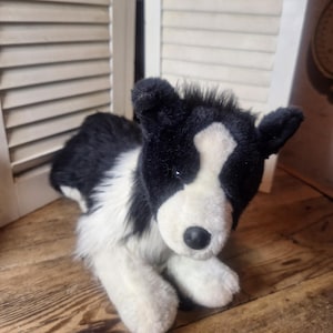 May include: A plush stuffed animal designed to resemble a border collie dog. The toy features soft black and white fur, a white blaze on its face, pointed ears, and a small black nose. It is depicted in a lying down position.