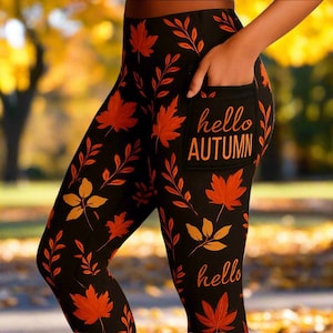 May include: Black leggings with an all-over pattern of orange and yellow autumn leaves and branches. The leggings have a pocket with the words "hello AUTUMN" printed in orange. The word "hello" is also printed on the leg.