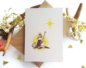 May include: A rectangular greeting card featuring a watercolor illustration of a shepherd holding a staff with two sheep standing on a patch of ground, illuminated by a bright yellow star above, all set against a textured off-white paper background.