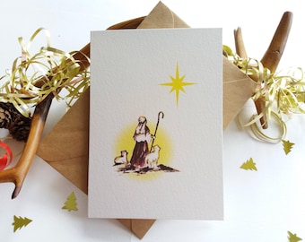 Puede incluir: Una tarjeta de Navidad con una estrella amarilla y un pastor con dos ovejas delante de él. El pastor lleva una túnica marrón y un bastón. La tarjeta está sobre un fondo blanco.
